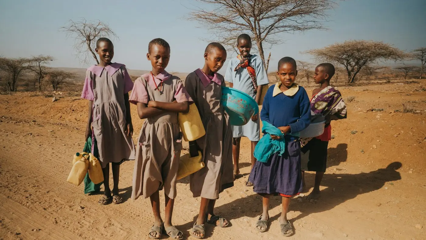 Six schoolchildren in uniform carrying water containers across dry red earth dotted with acacia trees